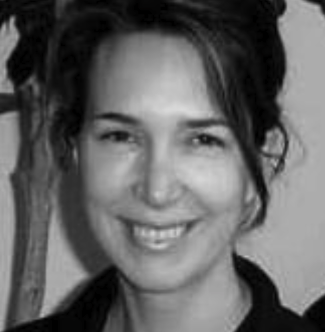 Black and white photo of a woman smiling. She has dark hair pulled back and is wearing a black top. The background shows part of a plant and a light-colored wall.